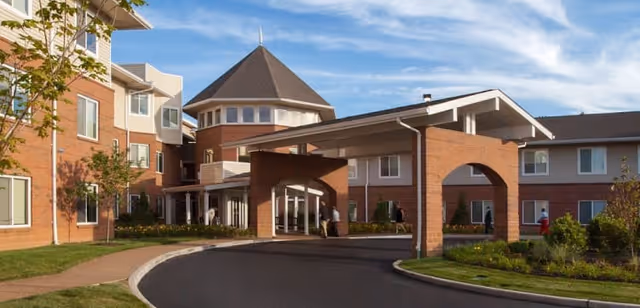 Exterior view of a senior living facility named Anna House, featuring a brick building with multiple windows, a covered entrance with arches, a driveway, and landscaped greenery under a partly cloudy sky.