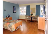 A well-lit bedroom in a senior living facility with a single bed covered in a floral quilt, a small wooden table with two chairs near a window with blue curtains, a wooden dresser with a TV on top, and a framed picture on the wall.