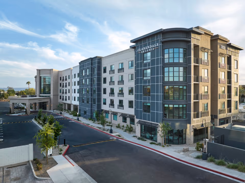 Exterior view of Clarendale Arcadia, a modern multi-story senior living facility with large windows and a covered entrance. The building is surrounded by a parking lot and landscaped areas with small trees and shrubs under a partly cloudy sky.