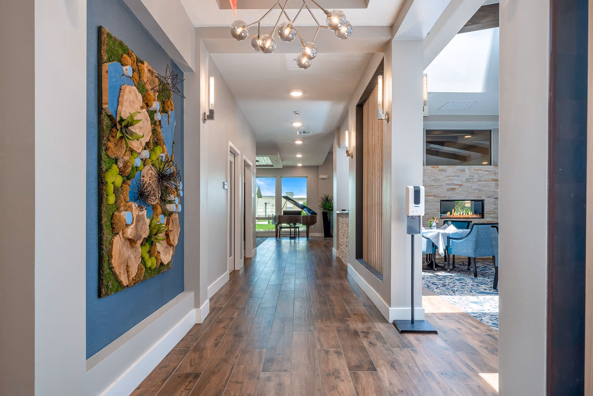 A bright hallway with wooden flooring and modern lighting fixtures on the ceiling. On the left wall, there is a colorful textured art piece with natural elements like wood slices and moss. At the end of the hallway, a grand piano is visible near large windows showing an outdoor view. To the right, there is a glimpse of a cozy dining area with blue chairs, a table set with a white tablecloth, and a modern fireplace with a stone surround.