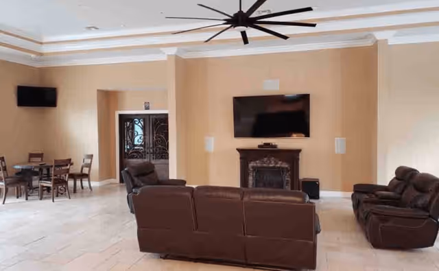 A spacious living room area with beige walls and tiled floor. The room features a large brown leather sofa set, a flat-screen TV mounted above a decorative fireplace, a ceiling fan with multiple blades, and a small dining table with chairs in the corner near a set of ornate double doors.