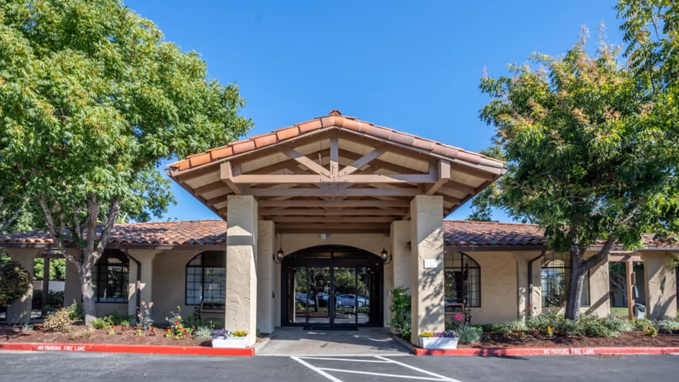 Front exterior view of a single-story building with a tiled roof and a covered entrance supported by columns, surrounded by trees and landscaping under a clear blue sky.