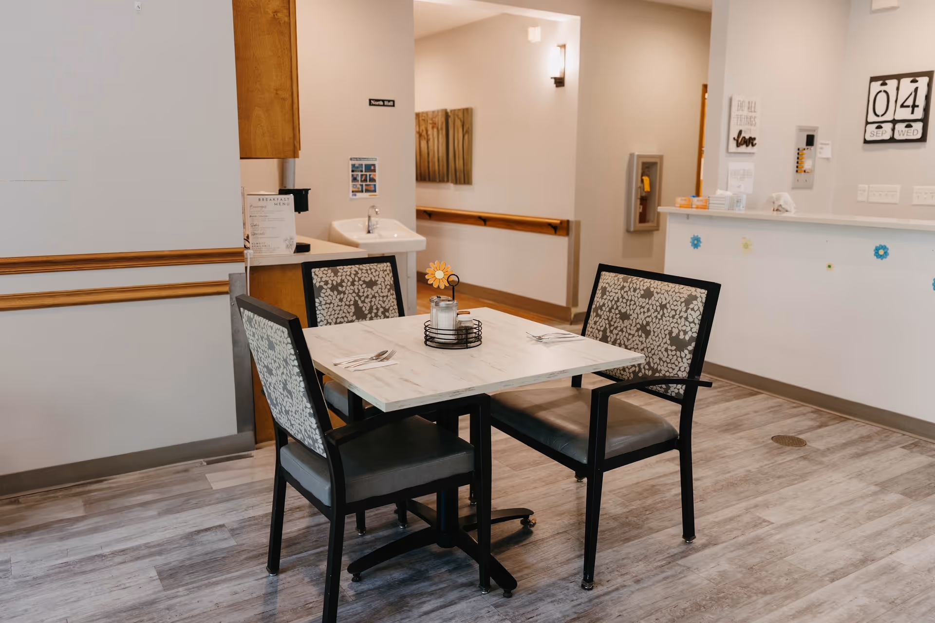 A small dining area in an assisted living facility with a square table set for four, featuring floral-patterned chairs. The room has light-colored walls, wood trim, and a light wood floor. In the background, there is a hallway, a wall-mounted sink, a bulletin board, and a reception counter with decorative items and a calendar showing September 4, Wednesday.