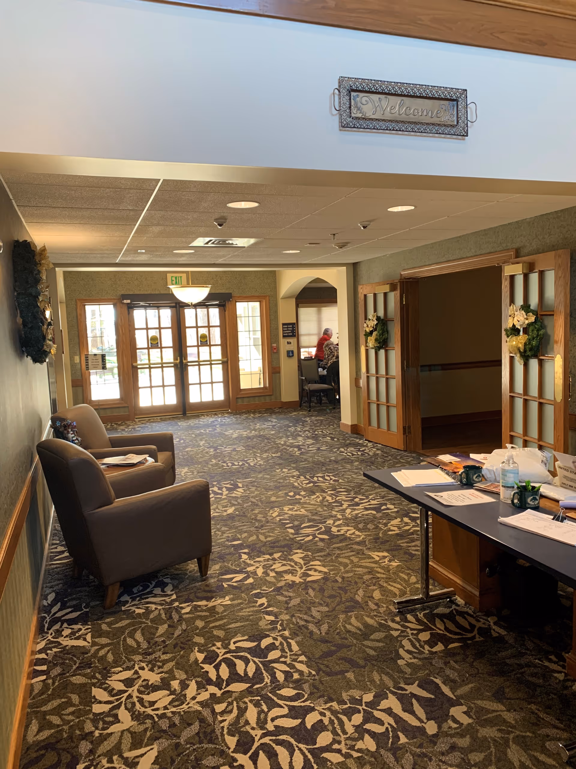 Carpeted lobby hallway of a senior living community with chairs, a table with papers, and glass double doors under a 'Welcome' sign.