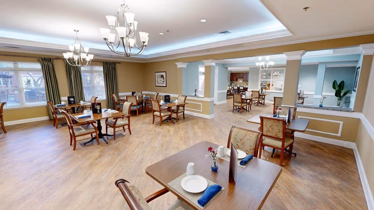 A spacious dining room in Dunwoody Place with multiple wooden tables and cushioned chairs arranged neatly. Each table is set with white plates, blue napkins, and small flower vases. The room has large windows with green curtains, beige walls, wooden flooring, and elegant chandeliers hanging from the ceiling.