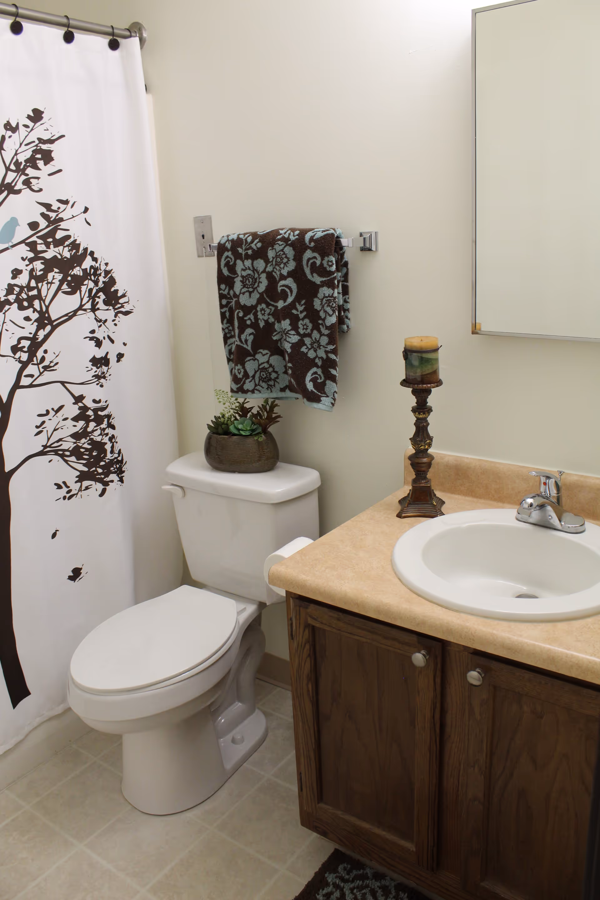 A clean bathroom featuring a white toilet with a small potted plant on the tank, a towel rack with a dark brown and light blue floral towel, a beige countertop with a white sink, a decorative candle holder with a candle, a mirror above the sink, and a shower curtain with a tree and bird design.