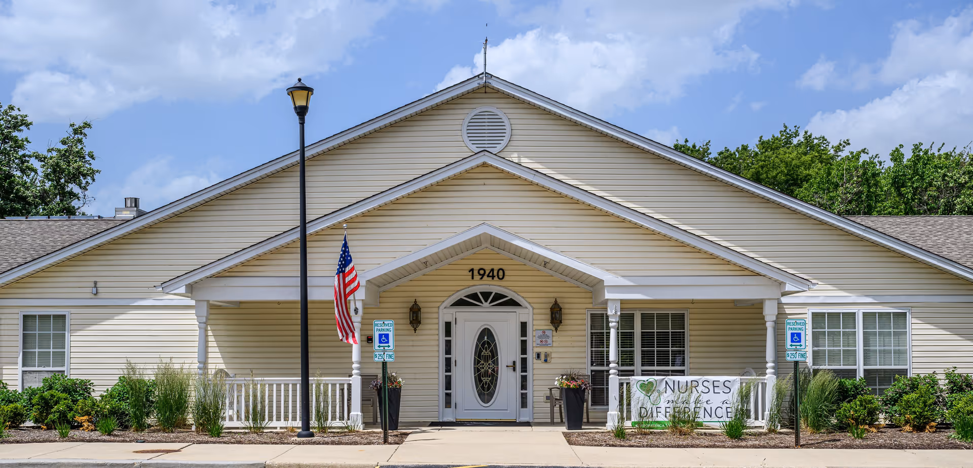 Front exterior view of a single-story beige building with white trim, a central entrance with a white door featuring decorative glass, and the number 1940 above it. There is an American flag on a pole near the entrance, two handicap parking signs, and a banner that reads 'Nurses make a difference' hanging on the porch railing. The building is surrounded by greenery and bushes under a partly cloudy blue sky.