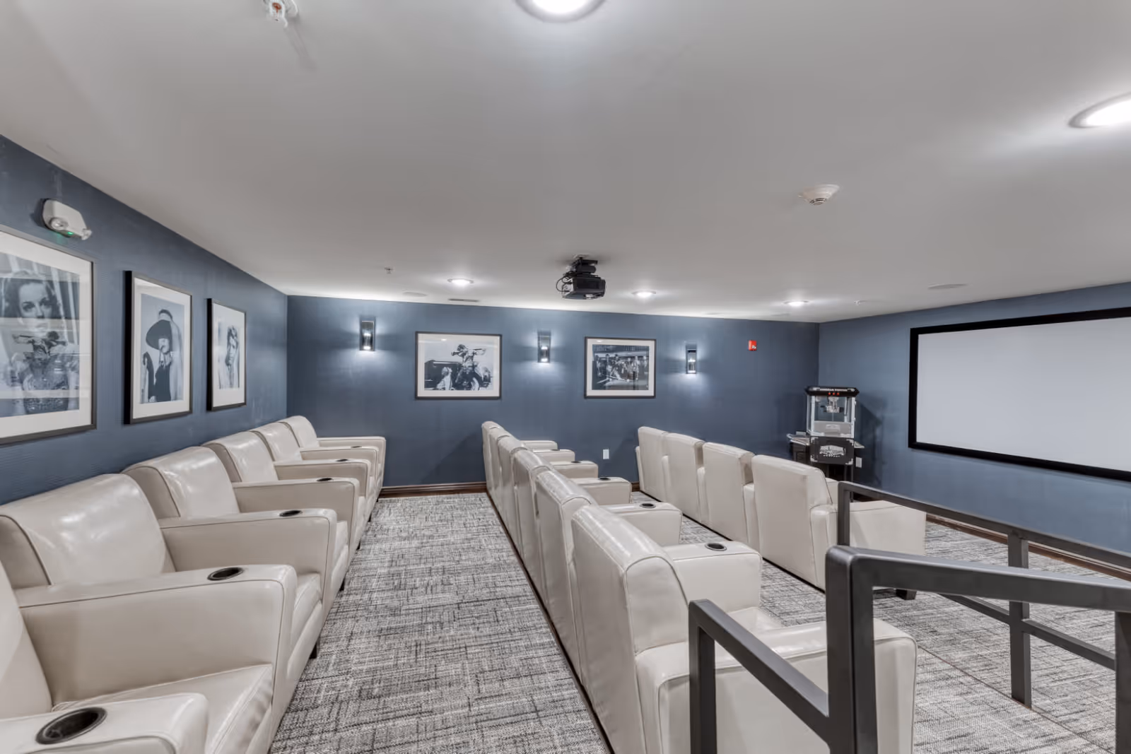 A senior living facility movie theater room with two rows of beige leather recliner chairs facing a large blank projector screen. The walls are painted blue and decorated with black and white framed photographs. A popcorn machine is visible in the corner of the room.