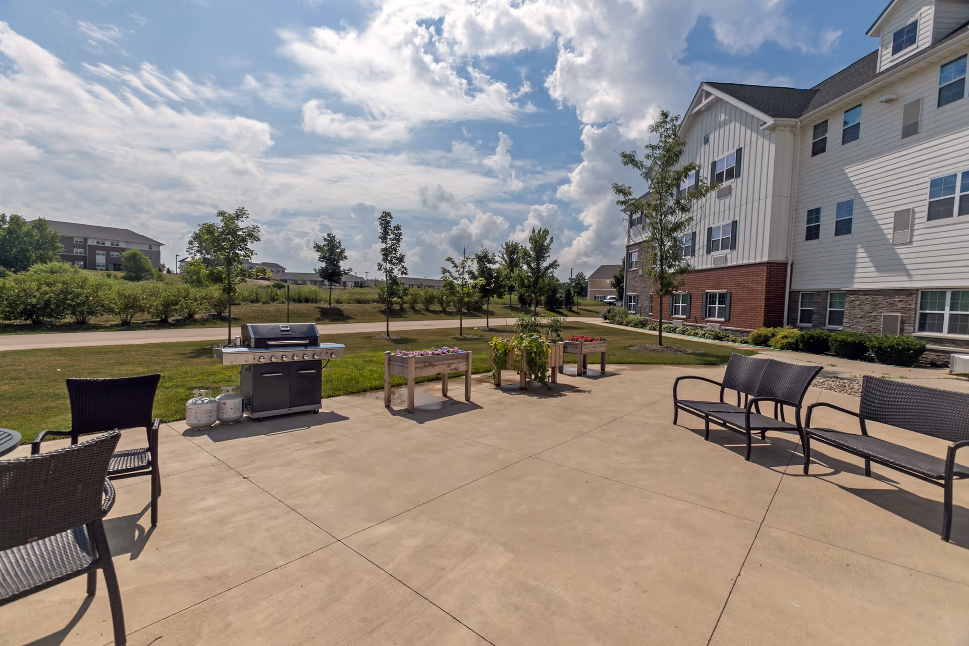 Outdoor patio area with a barbecue grill, planters and several benches beside a multi-story building under a partly cloudy sky.