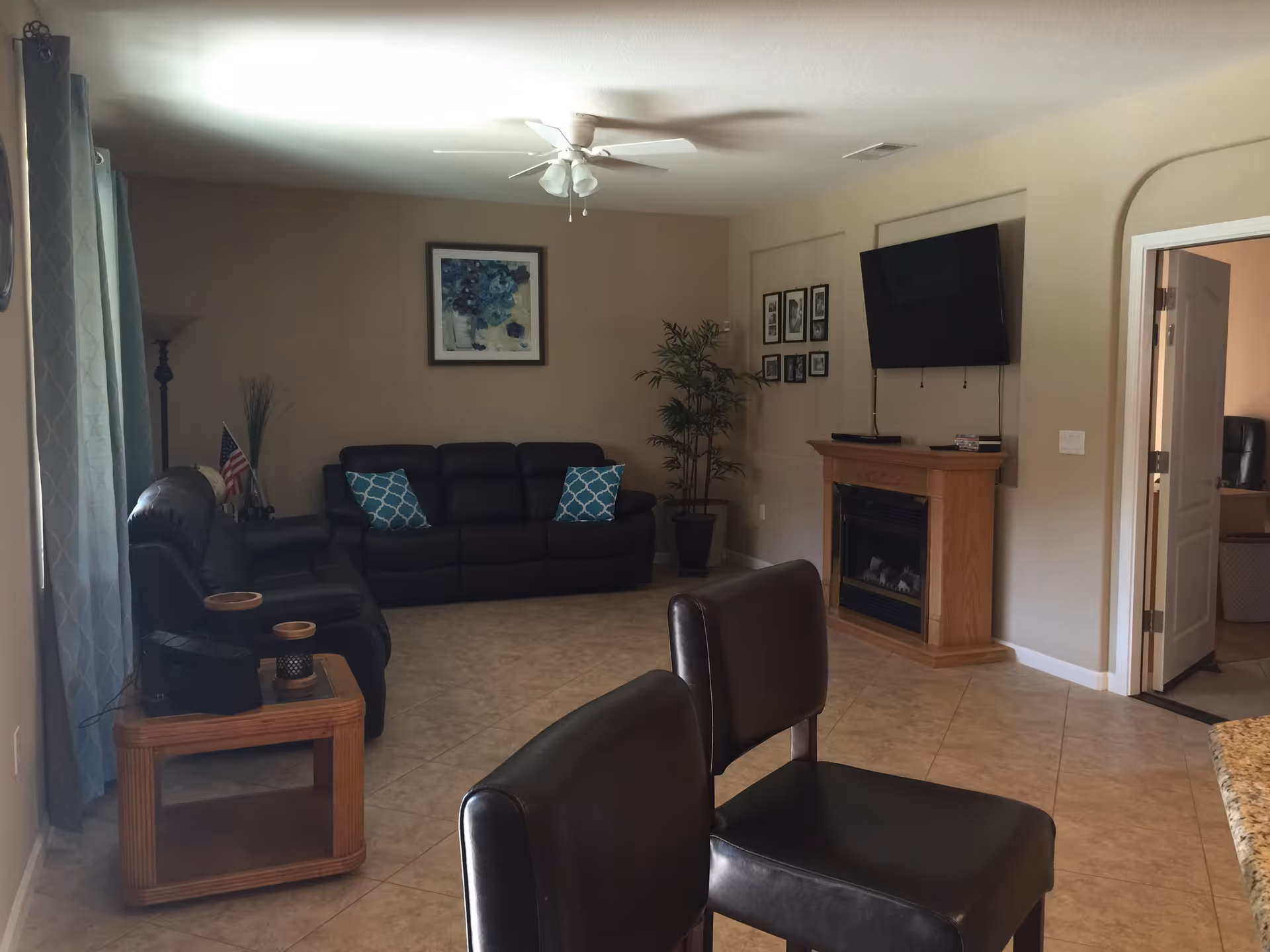 Interior view of a living room with beige walls and tiled floor. The room features two black leather sofas with blue patterned cushions, a wooden side table with decorative items, a ceiling fan with lights, a wall-mounted flat-screen TV above a wooden fireplace, framed pictures on the walls, a tall potted plant, and an open door leading to another room.