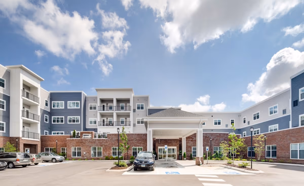 Front exterior of a modern multi-story senior living building with a covered entrance, balconies and a parking lot under a blue sky.