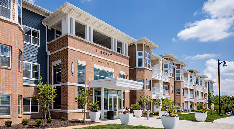 Exterior front entrance of the Liberty at Shoal Creek building with brick facade, covered entry, balconies, and landscaped planters under a blue sky.