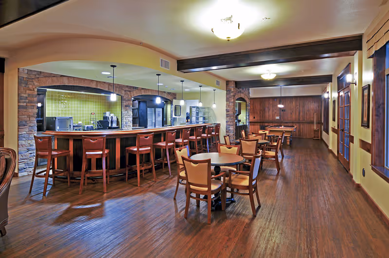 Spacious warm-toned dining area with a long bar and high stools, several round tables and chairs, and wood flooring.