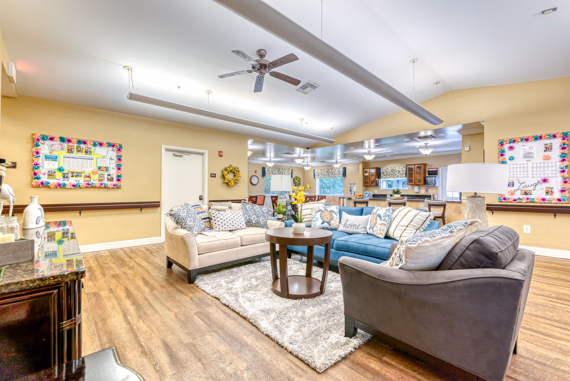 A spacious and well-lit common living area in a senior living facility featuring three sofas arranged around a round wooden coffee table on a light-colored rug. The room has wooden flooring, beige walls, ceiling fans, and overhead lighting. In the background, there is a kitchen area with wooden cabinets, a countertop, and bar stools. Colorful bulletin boards with decorations are mounted on the walls.