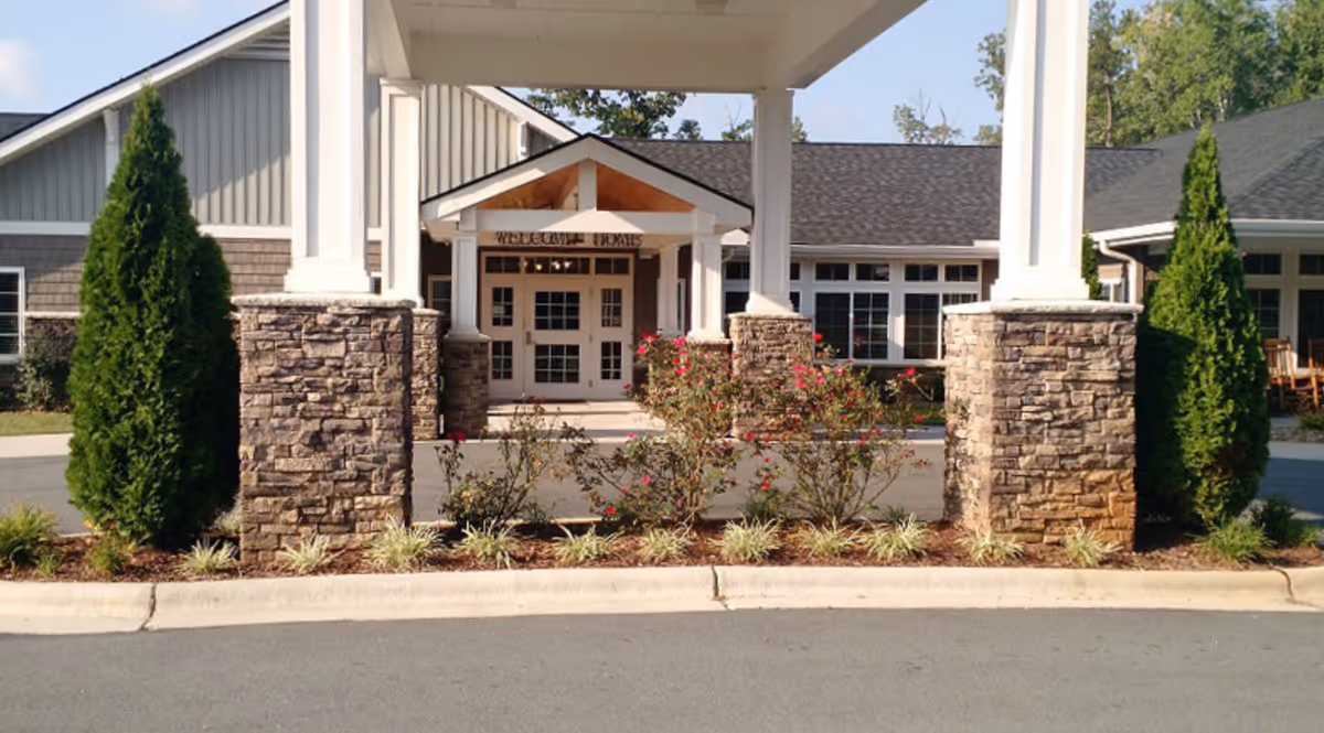 Entrance of Alamance House facility showing a covered driveway with stone pillars, landscaped bushes and trees, and a building with large windows and a peaked roof in the background.