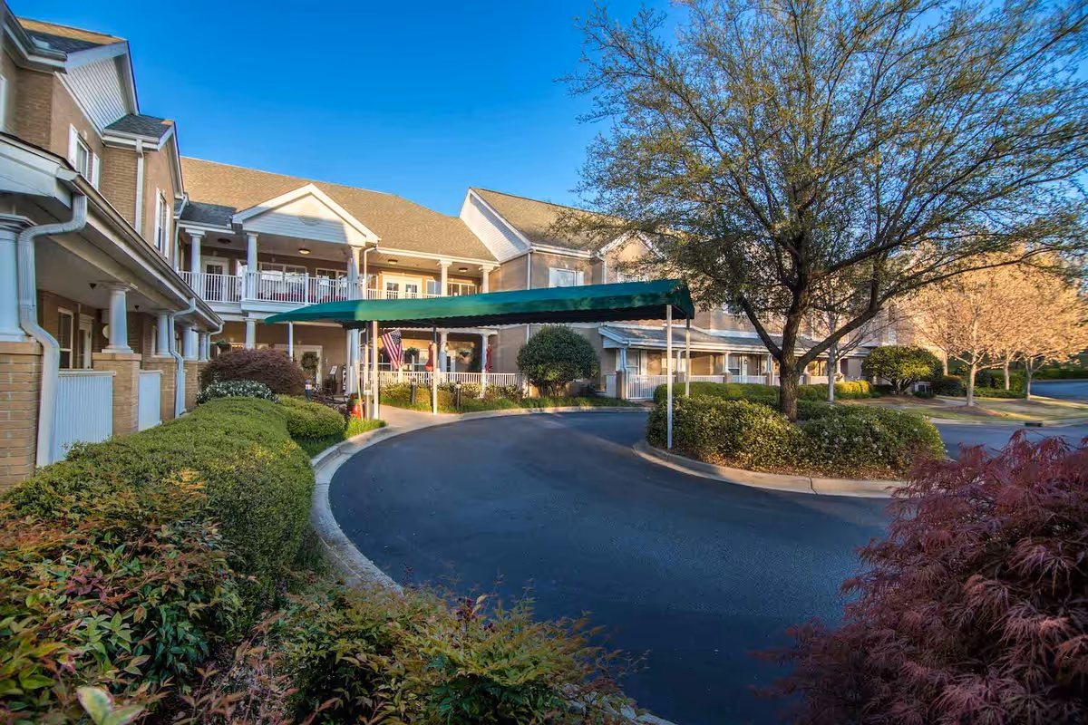 Exterior view of a senior living facility named The Gardens at Eastside, showing a two-story building with balconies, a covered driveway entrance, well-maintained bushes, trees, and a clear blue sky.