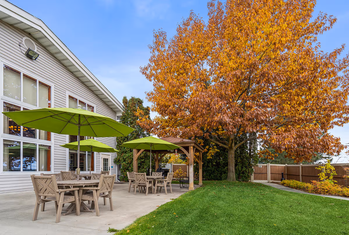 Outdoor patio area at a senior living facility with several tables and chairs under green umbrellas, a large tree with orange autumn leaves, a wooden pergola, and a well-maintained lawn and garden area.