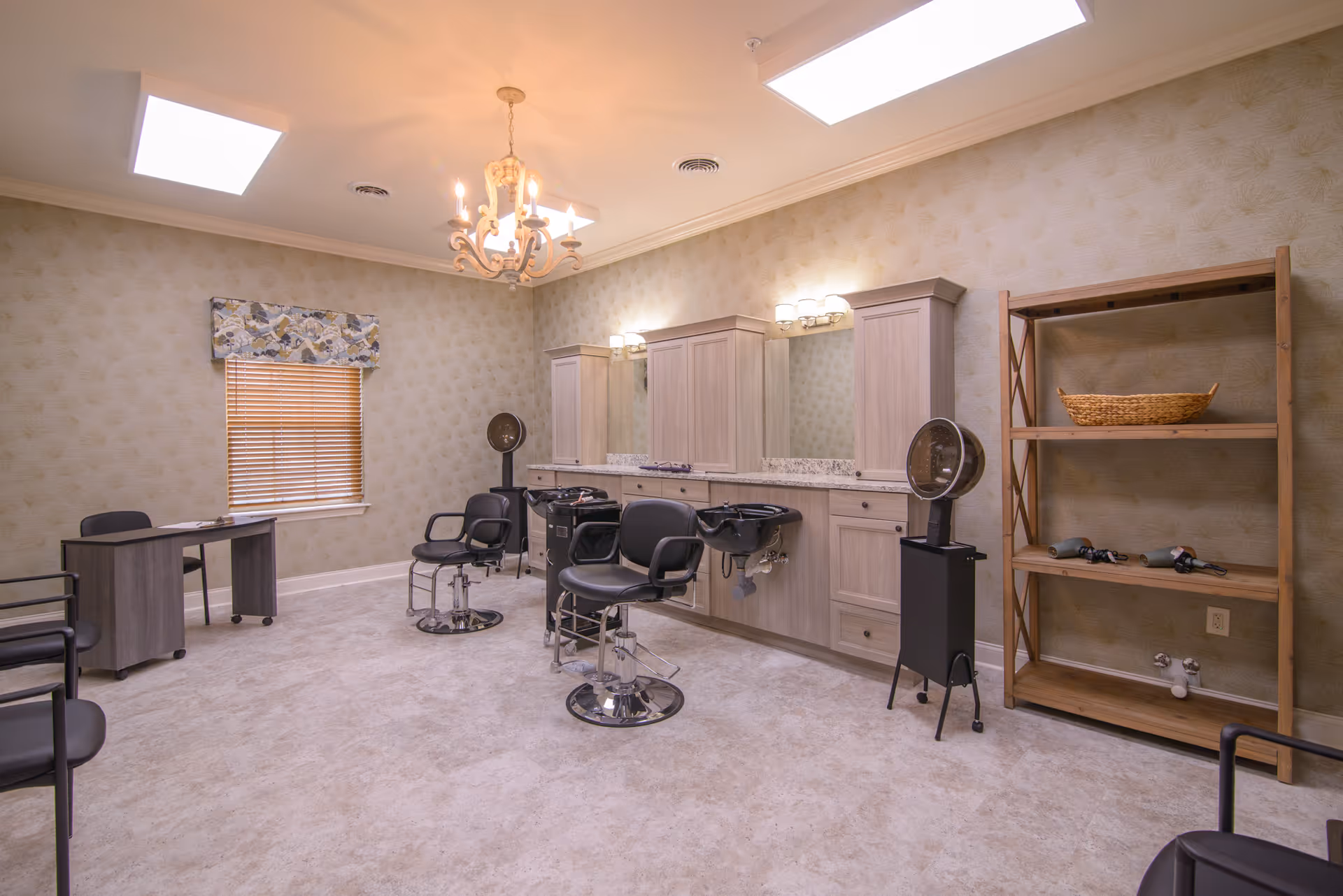 A spacious hair salon room with three black salon chairs in front of a long counter with sinks and mirrors. The room has beige patterned wallpaper, a window with blinds and a floral valance, a chandelier, and a wooden shelving unit holding a basket and hair dryers. There is also a small desk and chairs in the corner.