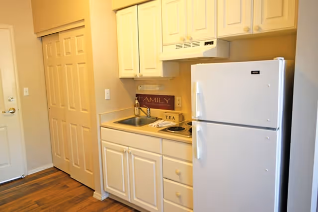 Small kitchen area with white cabinets, a white refrigerator, a sink, and a countertop with a small stove. There is a wooden floor and a door with a closet next to it.
