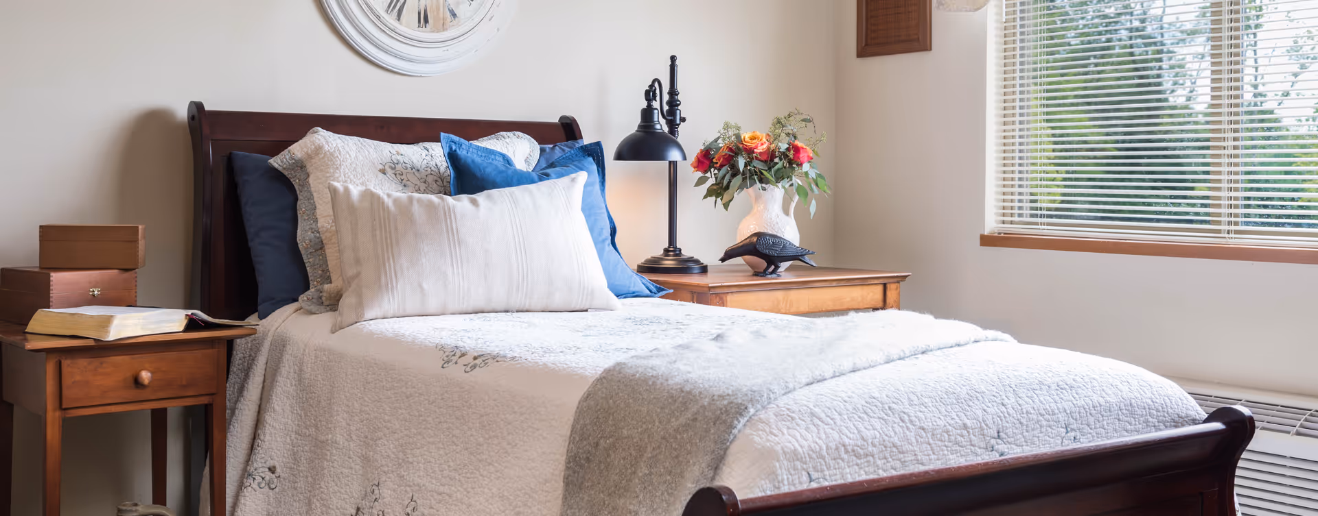 Sunlit bedroom with a neatly made wooden bed, layered pillows and a throw, wooden bedside tables holding a lamp, books and a vase of flowers beside a window with blinds.