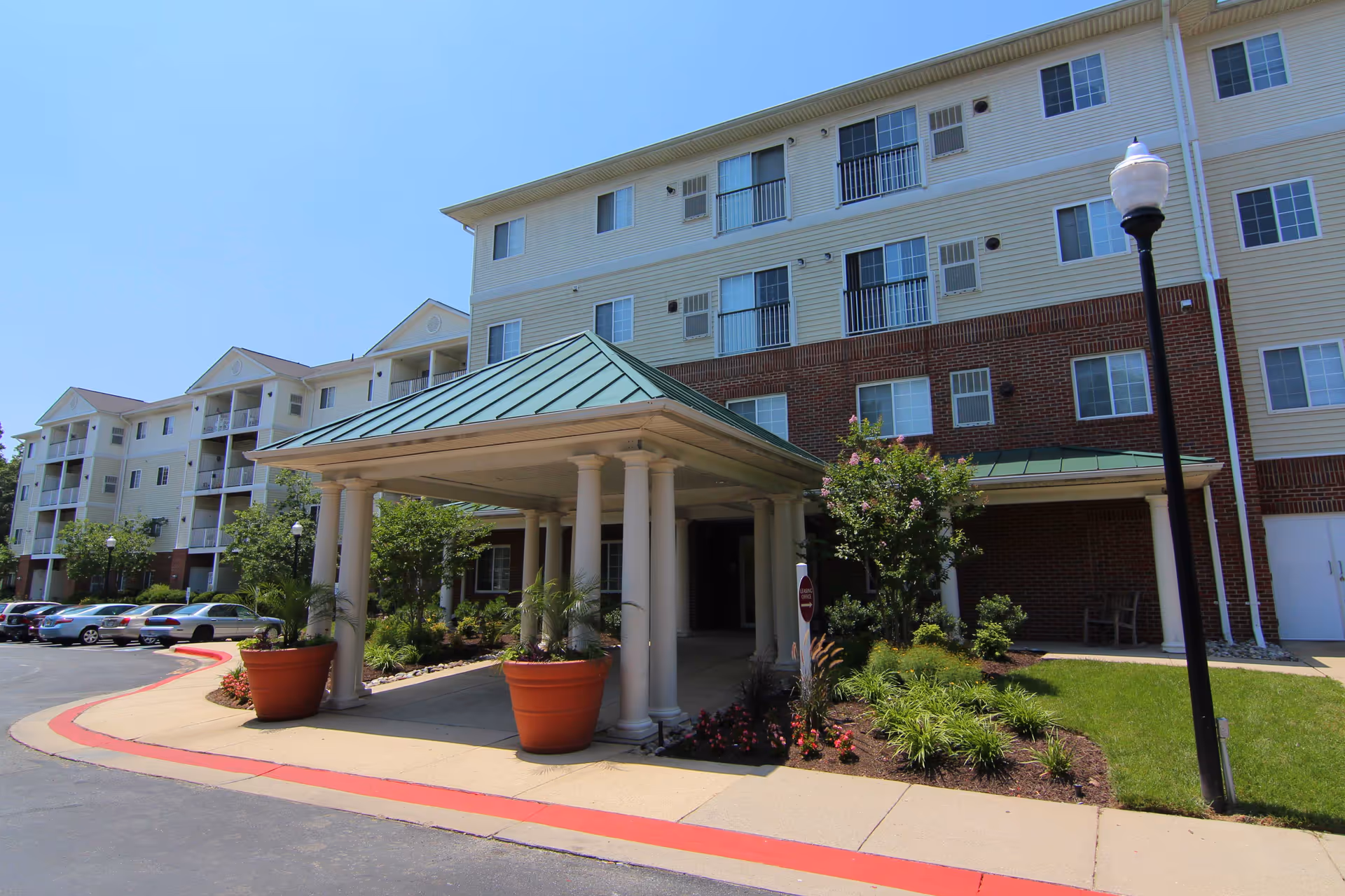 Exterior view of a multi-story senior living facility named Glen Forest with a covered entrance supported by columns, large potted plants, landscaped greenery, and a parking area with several cars under a clear blue sky.