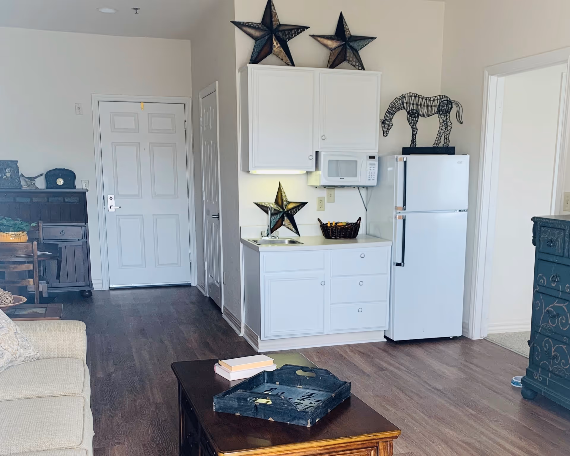 Open studio living area showing a small kitchenette with white cabinets, microwave and refrigerator, a coffee table and couch, and decorative star and horse accents on the wall.