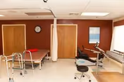 A physical therapy or rehabilitation room with exercise equipment including a treadmill, parallel bars, a walker, and a padded therapy table. The room has brown walls, two wooden doors, a clock, and a window with white curtains allowing natural light.