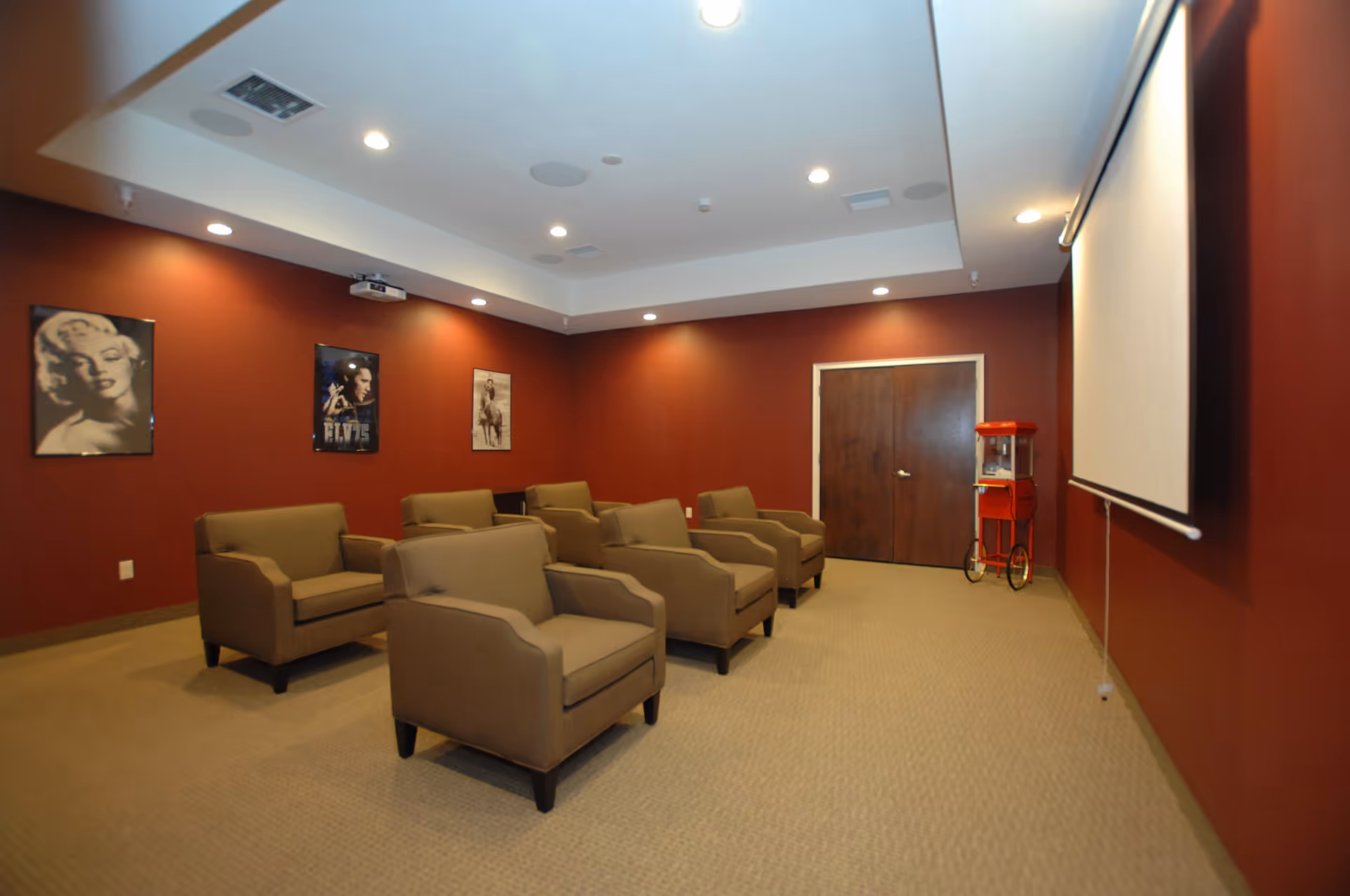 A small theater room with six beige armchairs arranged in two rows facing a large white projection screen. The walls are painted dark red and decorated with framed posters of Marilyn Monroe, Elvis Presley, and a cowboy. A red popcorn machine on wheels is positioned near double wooden doors at the back of the room. The ceiling has recessed lighting and a mounted projector.