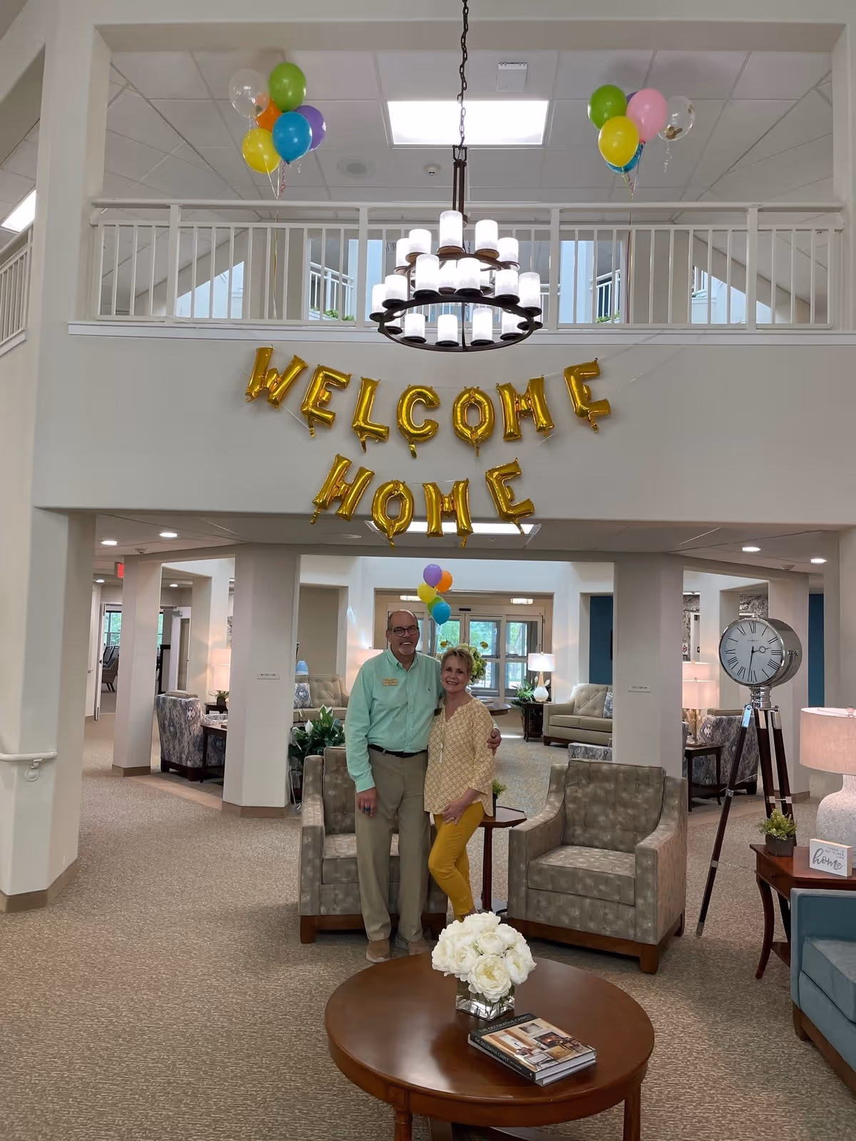 Interior lobby with gold 'WELCOME HOME' balloons, colorful balloons overhead, and two people posing between armchairs.