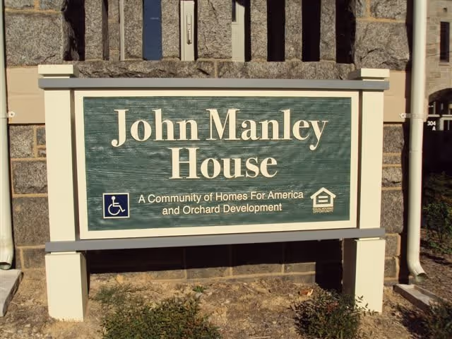 A green and white sign mounted on a stone wall that reads 'John Manley House, A Community of Homes For America and Orchard Development' with a wheelchair accessibility symbol and an equal housing opportunity logo.