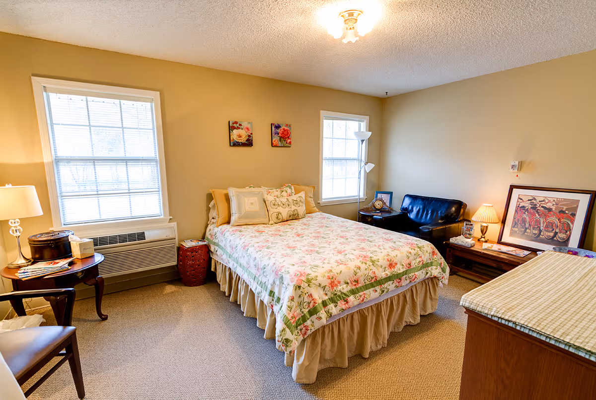 A cozy bedroom with a floral bedspread on a double bed, two windows letting in natural light, a wooden side table with a lamp and tissue box, a small black leather loveseat, a floor lamp, and framed artwork on the walls and a wooden dresser.