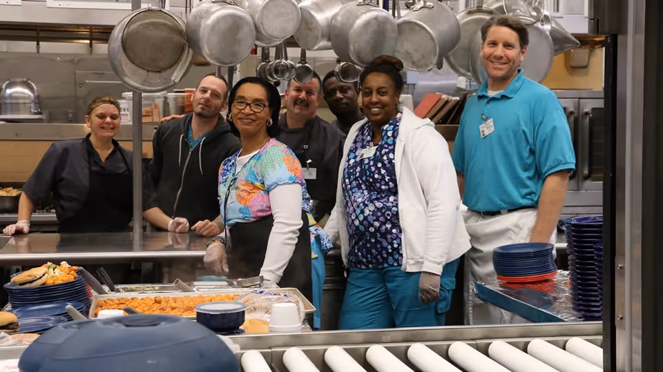 A group of six kitchen staff members standing together in a commercial kitchen behind a counter with trays of food and stacks of plates, smiling at the camera. Various pots and pans hang overhead.