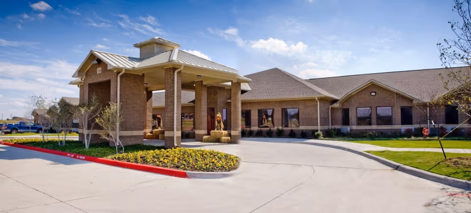 Exterior view of a single-story brick building with a covered entrance supported by columns, surrounded by landscaped greenery and a driveway under a partly cloudy sky.