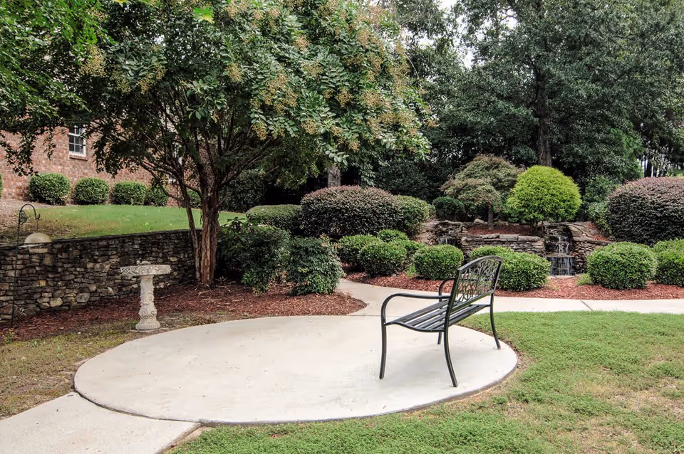 A peaceful outdoor garden area with a black metal bench on a concrete circular pad, surrounded by green grass, bushes, trees, and a small stone waterfall feature in the background.