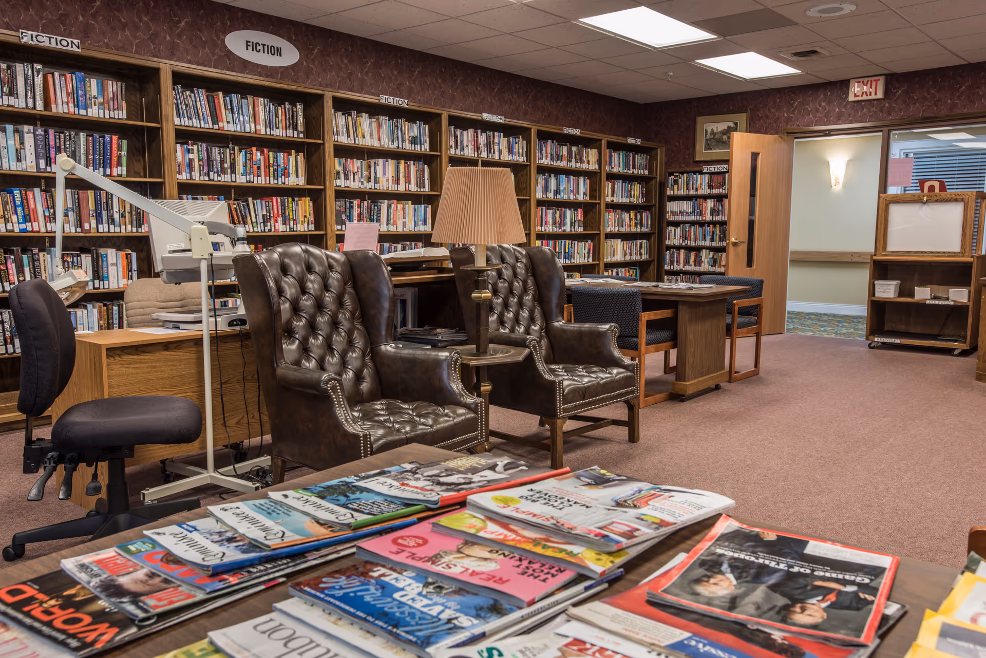 Interior view of a senior living facility library with shelves filled with fiction books, two brown leather tufted armchairs, a table with magazines in the foreground, and a desk with office chairs and a lamp. The room has carpeted floors and an open door leading to a hallway.
