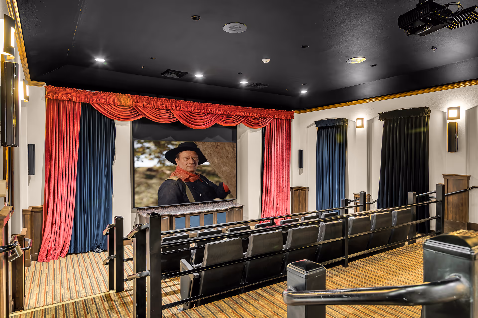 Interior view of a small theater room with tiered seating, black chairs, and a large screen showing a man in a cowboy hat and red scarf. The room features red and blue curtains around the screen, a black ceiling with recessed lighting, and striped carpet flooring.
