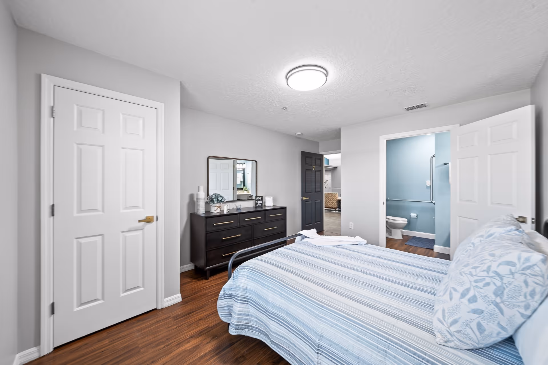 A bedroom with a bed covered in a blue and white striped bedspread and matching pillows. There is a dark wooden dresser with a mirror above it against one wall. The room has light gray walls and wood flooring. Two doors are visible: one closed white door and one open door leading to a bathroom with blue walls and a white toilet.
