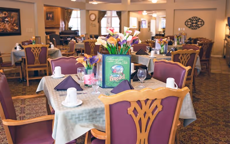 A dining room in a senior living facility with tables covered in beige tablecloths, set with white cups, glasses, and folded dark napkins. Each table has a centerpiece with colorful flowers and a decorative menu card. The room has wooden chairs with purple cushions and a patterned carpet.