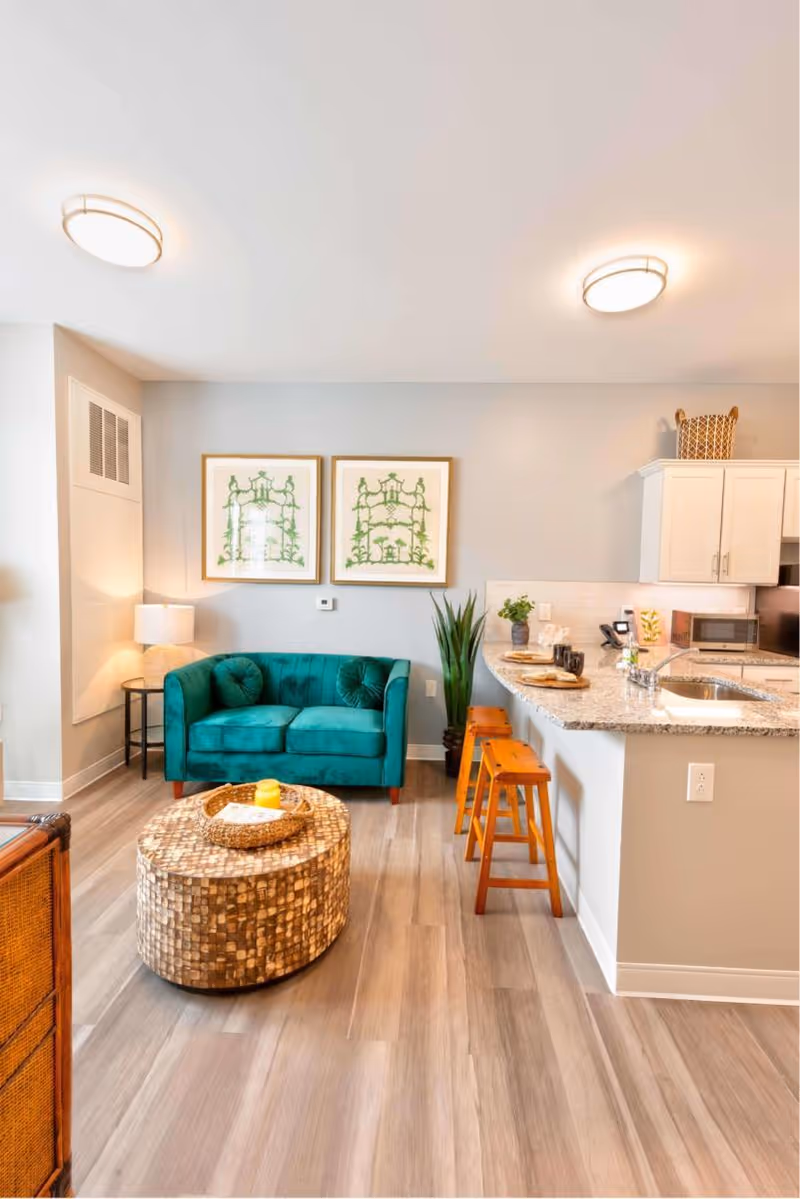 A bright and modern living area featuring a teal velvet loveseat with two round cushions, a round woven coffee table with a candle and a tray on top, and a kitchen counter with granite countertop and two wooden stools. The wall behind the loveseat has two framed botanical prints, and there is a tall green plant next to the counter. The floor is light wood, and the ceiling has two round light fixtures.
