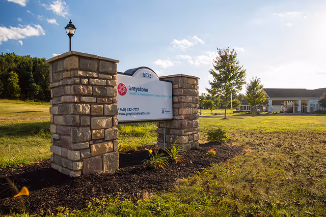 Stone pillars with a sign for Greystone Health & Rehabilitation Center at Cambridge, surrounded by grass and landscaping, with a building and trees in the background under a blue sky with some clouds.