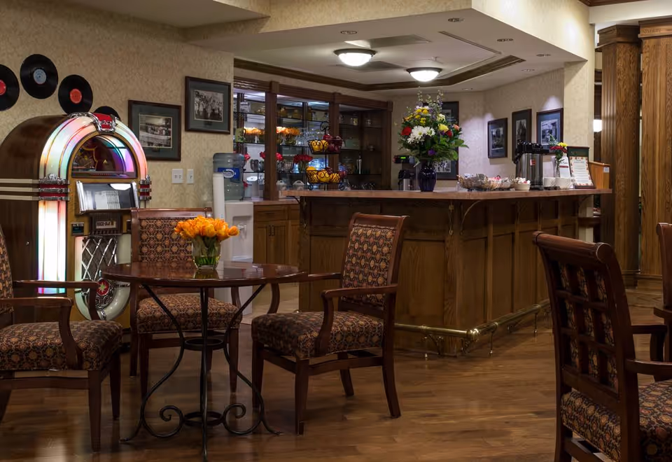 Cozy lounge/dining area with a wooden bar, patterned chairs around a small round table with orange tulips, and a vintage jukebox.