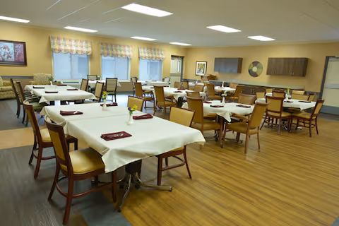 A spacious dining room with multiple tables covered in white tablecloths, each set with folded red napkins and small flower centerpieces. The room has wooden flooring, beige walls, large windows with valances, and several chairs arranged around the tables. There is wall art and a flat-screen TV mounted on the far wall.