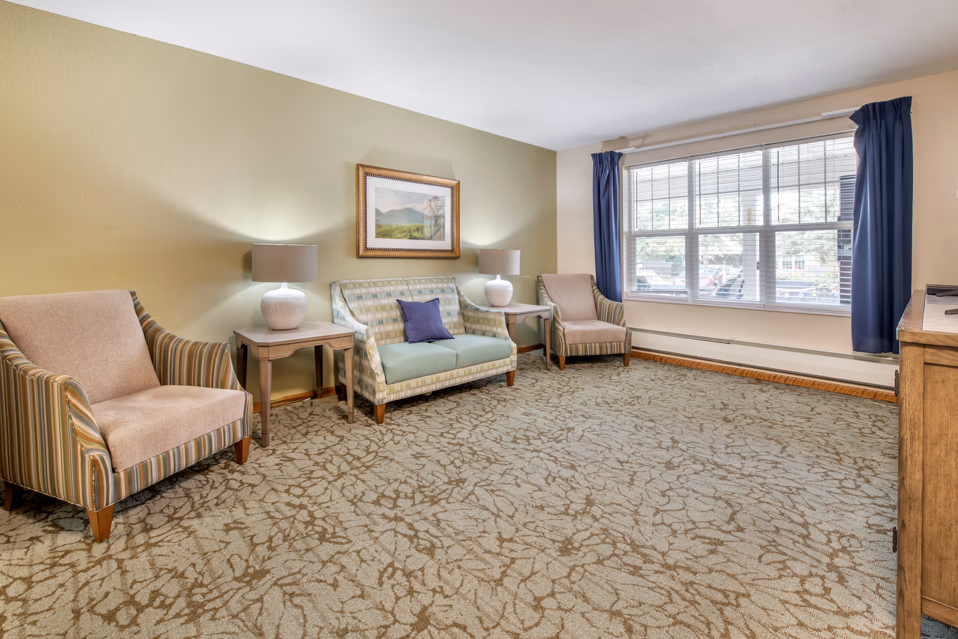 A cozy living room with two beige armchairs with striped sides, a patterned loveseat with a purple pillow, two side tables each with a white lamp, a framed landscape picture on a light green wall, a large window with blue curtains, and a patterned carpet.