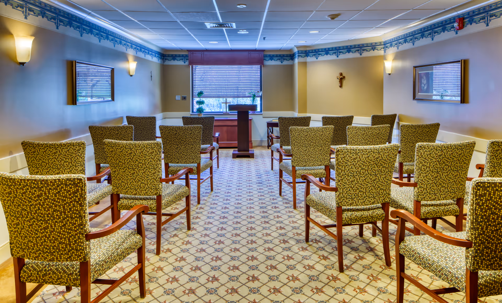 A small chapel or meeting room with rows of patterned upholstered chairs facing a wooden podium in front of a window with blinds. The room has beige walls with blue decorative trim near the ceiling, wall sconces for lighting, framed pictures on the walls, and a crucifix mounted on the right wall.