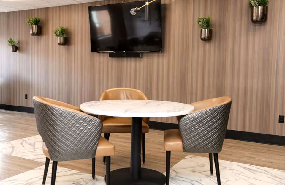 A modern seating area with a round marble table surrounded by three cushioned chairs with quilted backs. The background features a wood-paneled wall with a mounted flat-screen TV and several small potted plants in metallic wall planters.