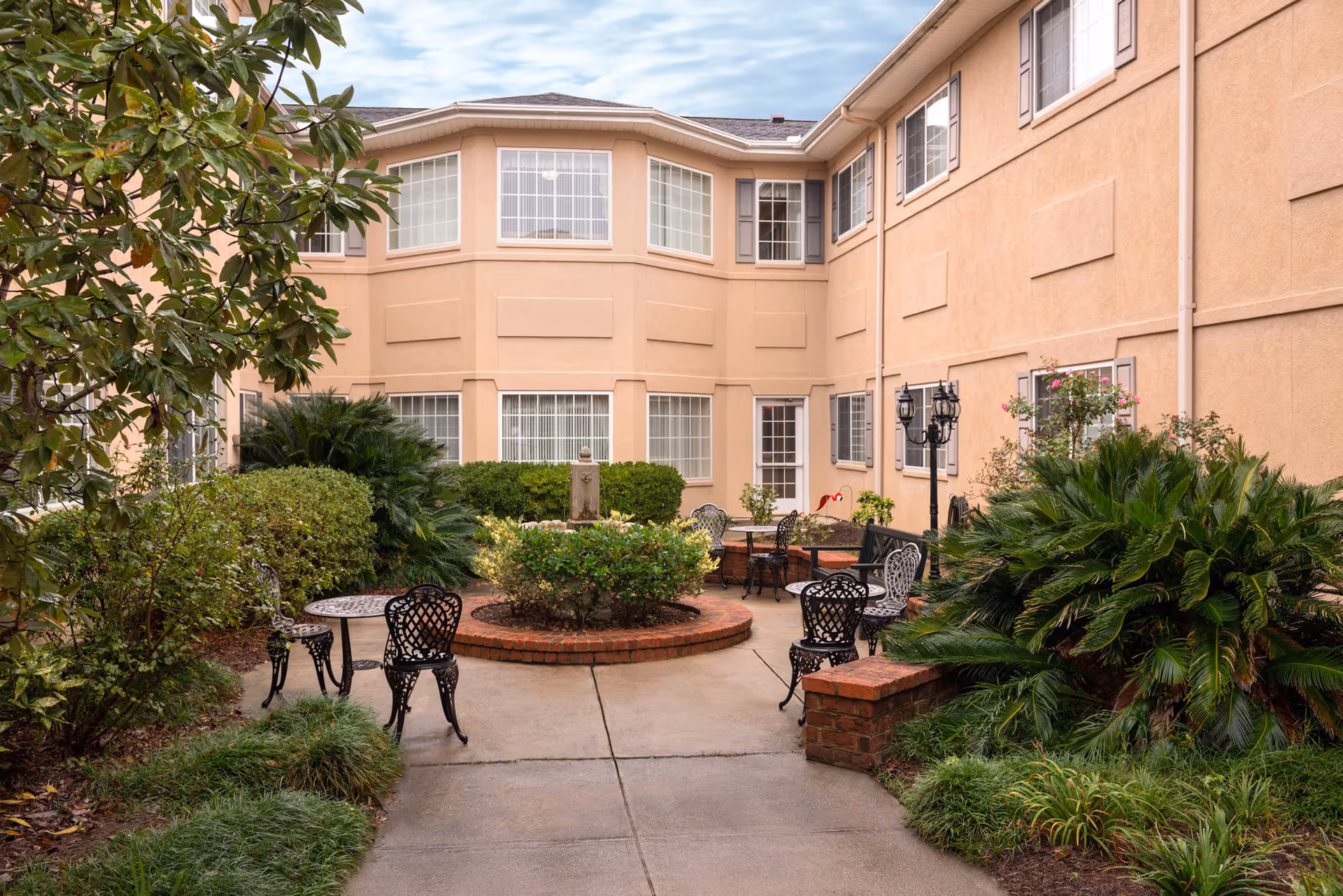 Courtyard patio with metal tables and chairs, a circular brick planter, shrubs, and the surrounding two-story building façade.