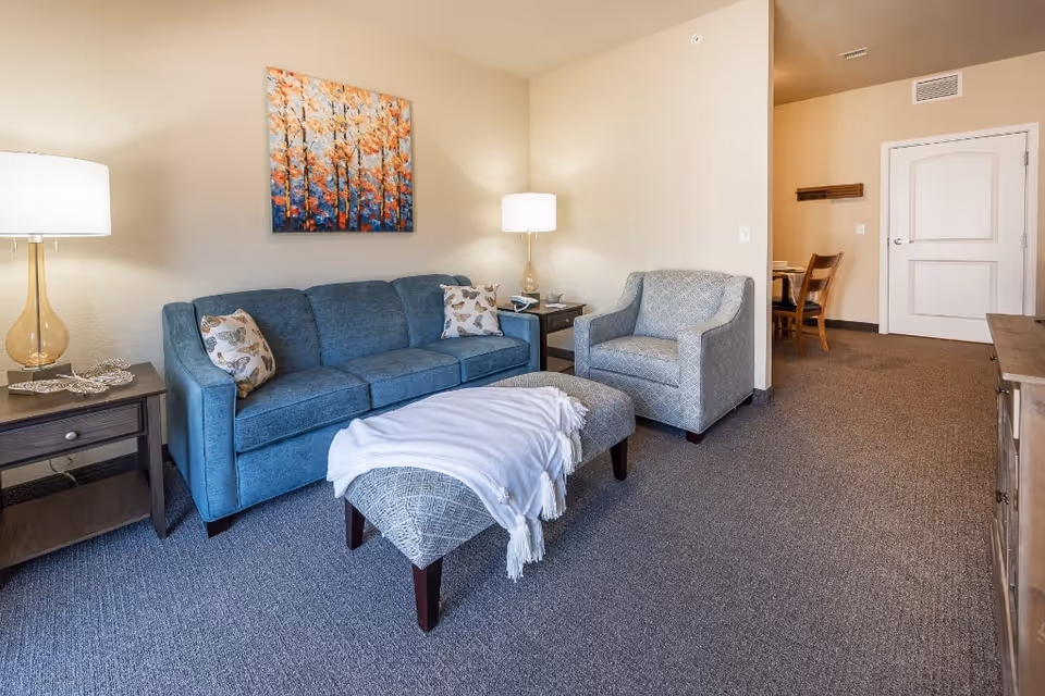 Furnished living room with a blue sofa, armchair, ottoman draped with a blanket, side tables, lamps, and wall art.