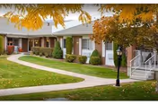 Exterior view of a single-story residential building with a light-colored roof and white walls, surrounded by neatly trimmed bushes and trees with autumn foliage. A paved walkway curves through a well-maintained lawn leading to the building entrance, which has a small porch with white railings and a black lamp post nearby.