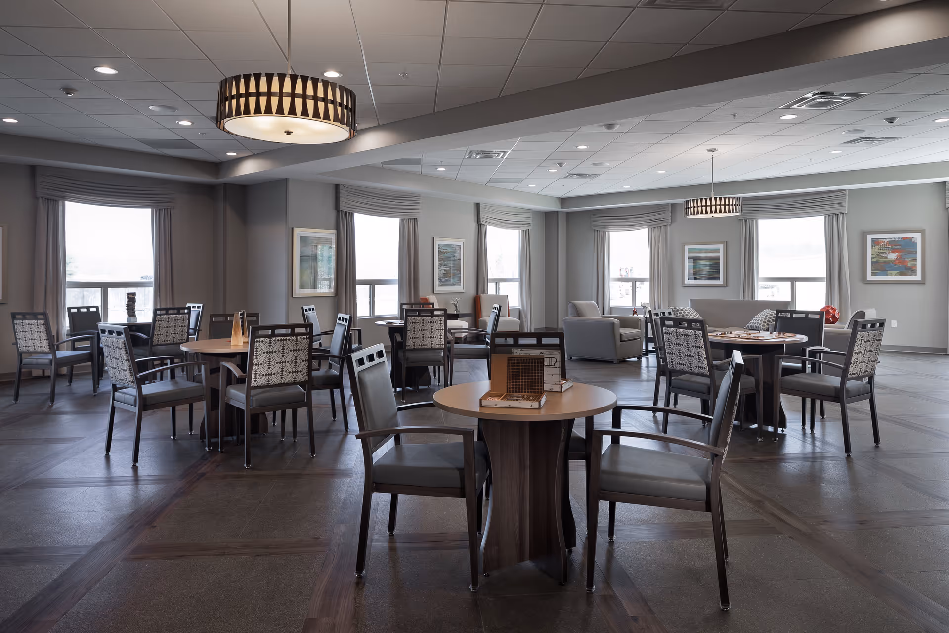 A spacious, well-lit common area with multiple round tables and chairs arranged for socializing or games. The room features large windows with curtains, modern ceiling lights, framed artwork on the walls, and a seating area with armchairs and a sofa in the background.