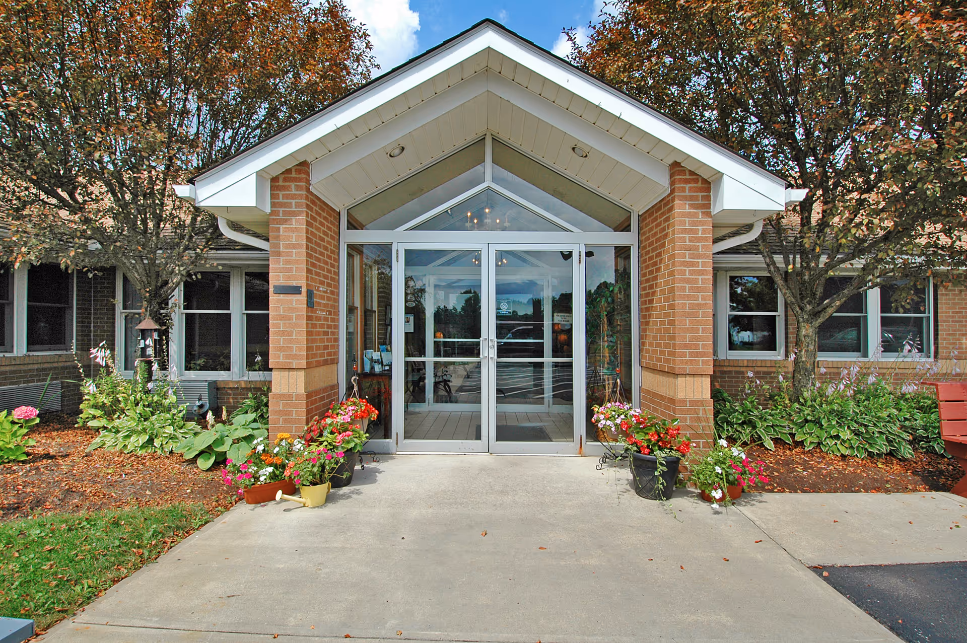 Entrance of a brick building with a glass double door under a white peaked roof. There are flower pots with colorful flowers on either side of the entrance and trees with autumn foliage flanking the building.